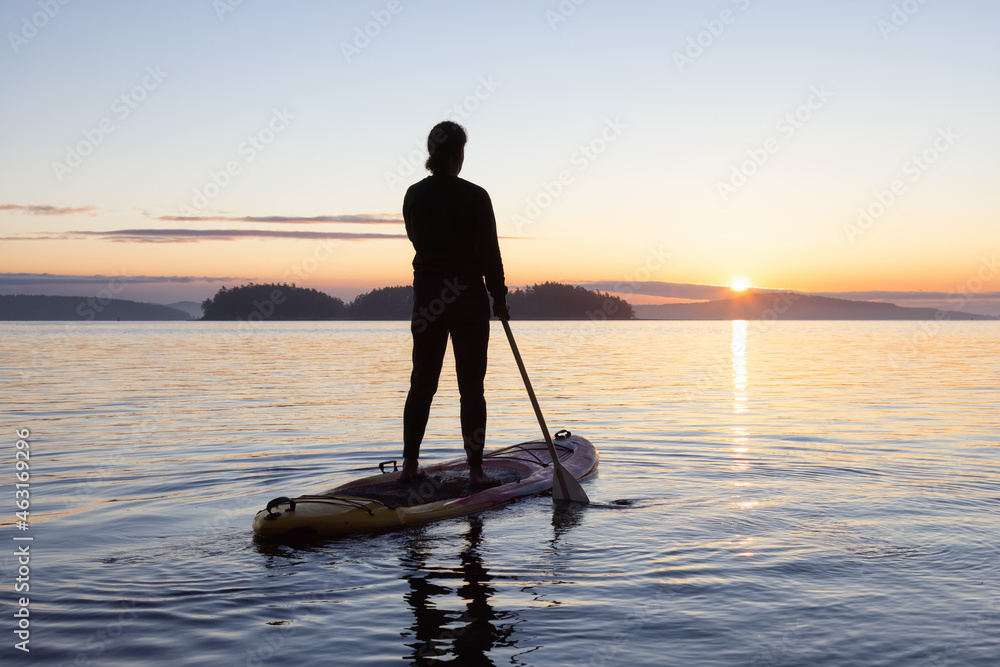 Adventurous Caucasian Adult Woman on a Stand Up Paddle Board is paddling on the West Coast of Pacific Ocean. Sunny Sunrise. Victoria, Vancouver Island, BC, Canada.