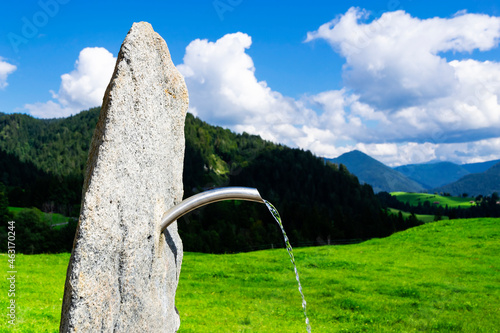 Bild auf Leinwand Jet of water of a spring flowing from a pipe attached to a large stone; meadow, mountains, blue cloudy sky in the background