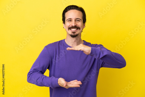 Young caucasian man isolated on yellow background holding copyspace imaginary on the palm to insert an ad