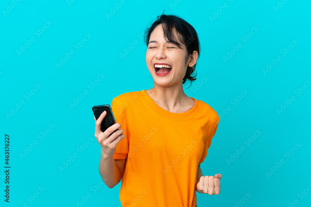 Young Vietnamese woman isolated on blue background using mobile phone and doing victory gesture Young Vietnamese woman isolated on blue background using mobile phone and doing victory gesture