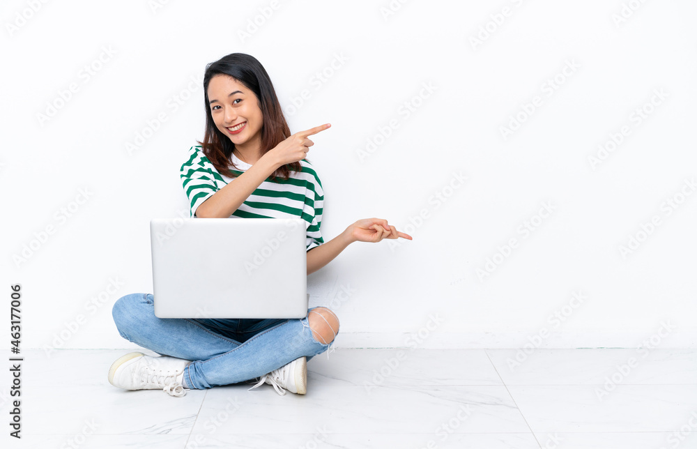 Young Vietnamese woman with a laptop sitting on the floor isolated on white wall pointing finger to the side and presenting a product Young Vietnamese woman with a laptop sitting on the floor isolated on white wall pointing finger to the side and presenting a product