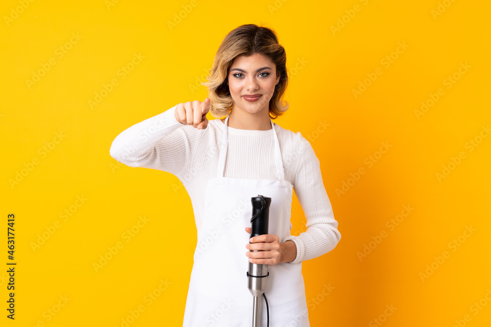 Girl using hand blender isolated on yellow background surprised and pointing front
