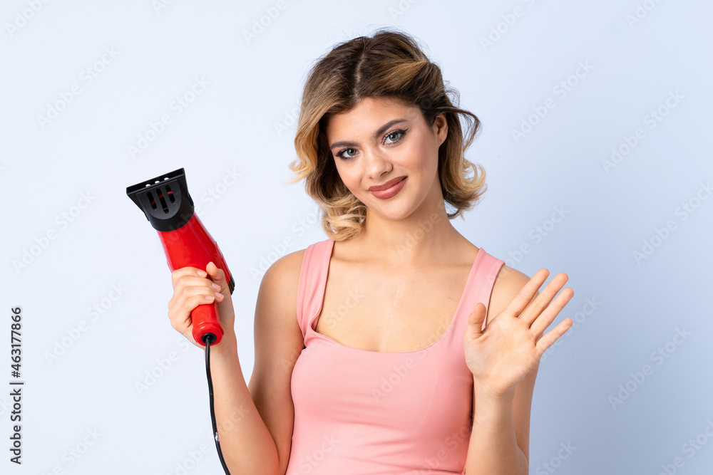 Russian girl holding a hairdryer isolated on blue background saluting with hand with happy expression Russian girl holding a hairdryer isolated on blue background saluting with hand with happy expression