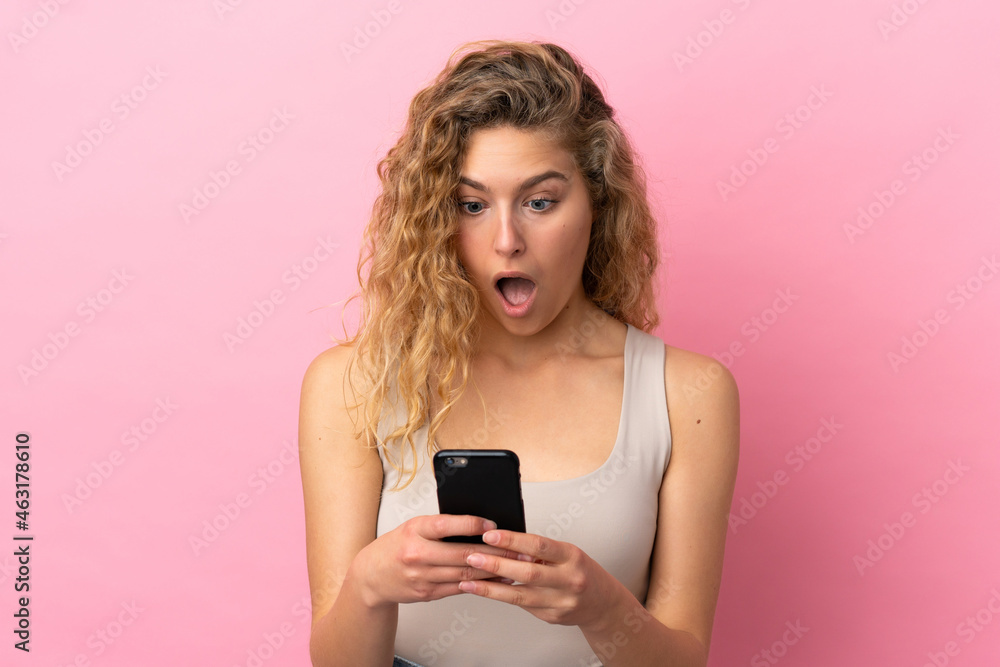 Young blonde woman isolated on pink background looking at the camera while using the mobile with surprised expression Young blonde woman isolated on pink background looking at the camera while using the mobile with surprised expression