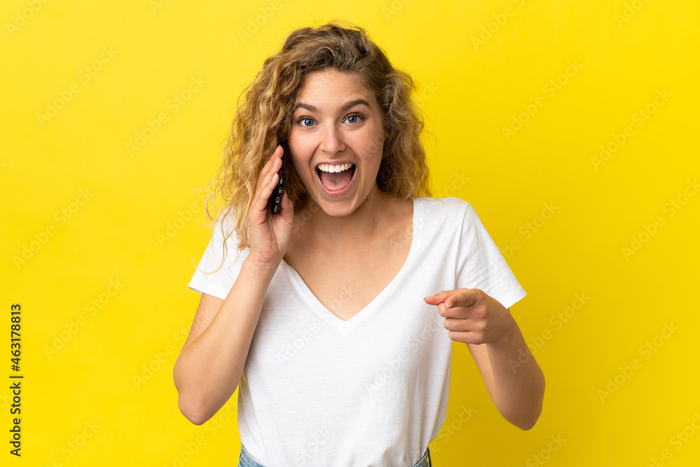 Young blonde woman using mobile phone isolated on yellow background surprised and pointing front Young blonde woman using mobile phone isolated on yellow background surprised and pointing front