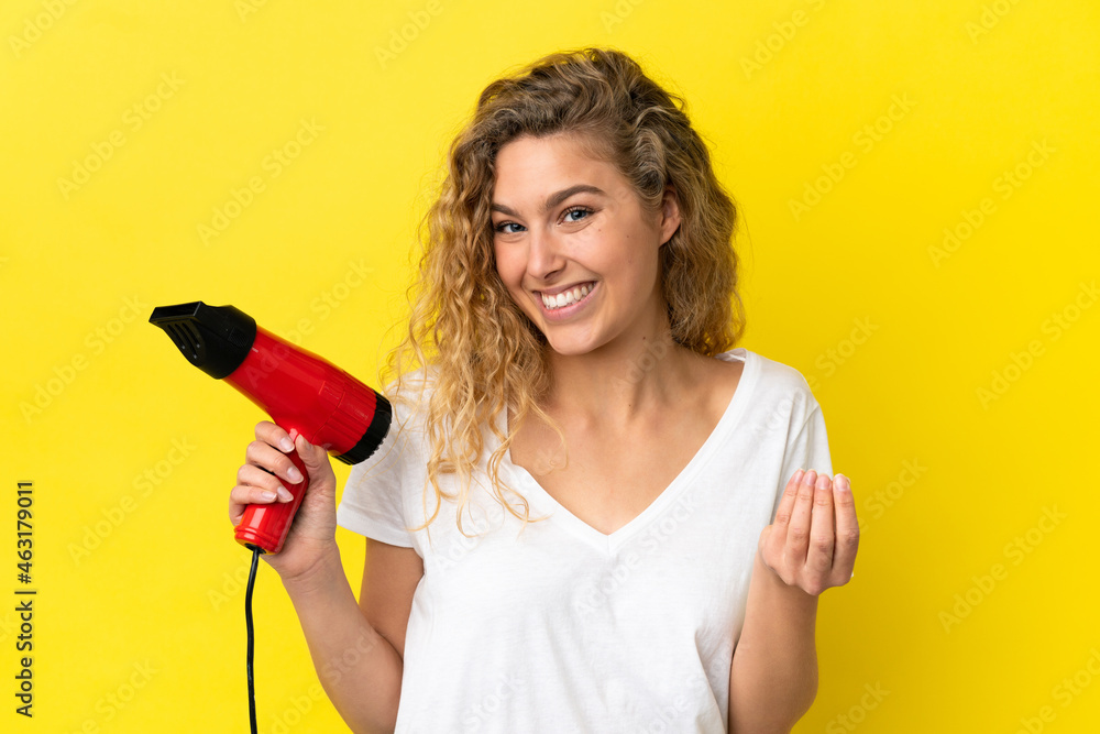 Young blonde woman holding a hairdryer isolated on yellow background making money gesture Young blonde woman holding a hairdryer isolated on yellow background making money gesture