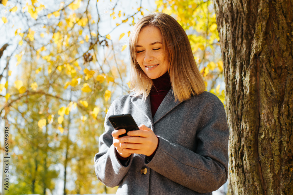 Happy young woman in gray coat using mobile phone, smiling looking at screen, browsing internet or social media, entertainment content outdoors. Selective focus on female face