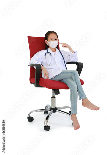 Asian little girl kid in doctor gown with pointing forefinger at medical mask sitting on chair isolated on white background. Image full length with Clipping path