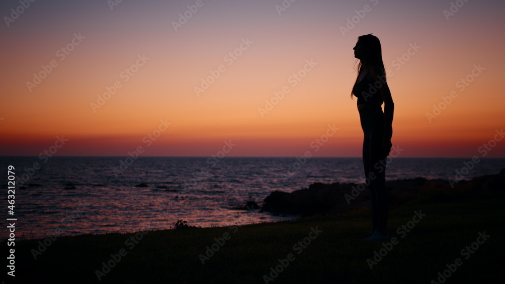 Fitness woman in silhouette doing squats exercises on beach near ocean. Young female in sportswear spending evening time for outdoors activity. Amazing colorful sunset on background. Fitness woman in silhouette doing squats exercises on beach near ocean. Young female in sportswear spending evening time for outdoors activity. Amazing colorful sunset on background.