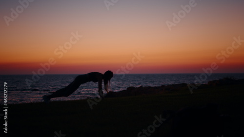 Strong young woman in silhouette standing in plank position during workout on ocean beach. Female athlete in sport outfit training abs with colorful summer sunset on background.