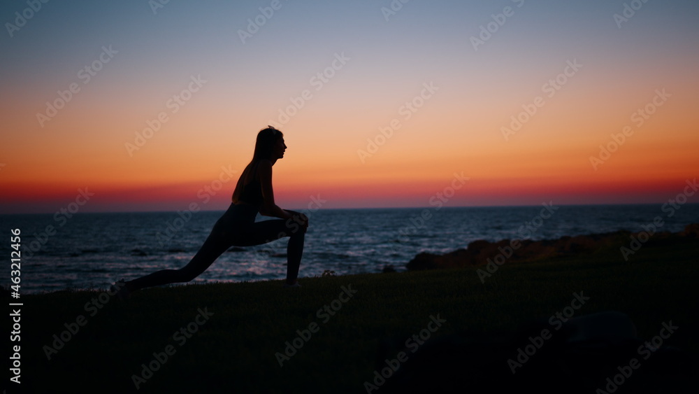 Silhouette of fitness young woman stretching legs after active workout on beach. Beautiful summer sunset over ocean. Concept of healthy and active lifestyles.
