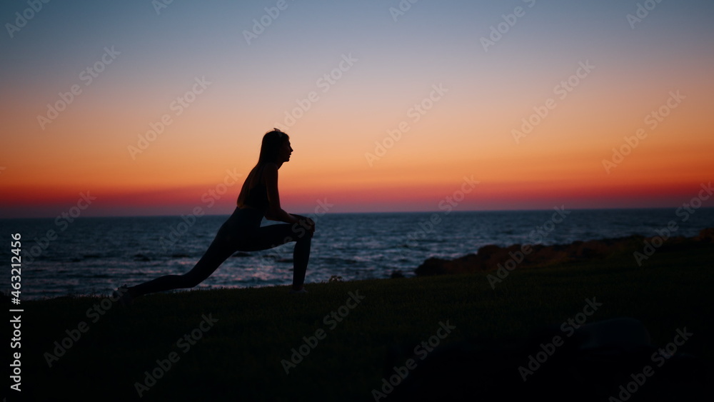 Fototapeta premium Silhouette of fitness young woman stretching legs after active workout on beach. Beautiful summer sunset over ocean. Concept of healthy and active lifestyles.