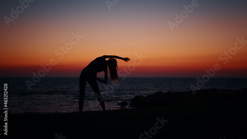 Young woman with slender body practising in fitness exercises during evening time near ocean. Silhouette of active healthy female. Regular training for body care.