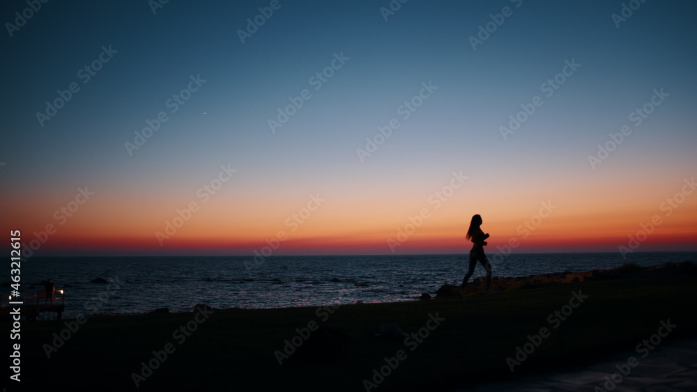 Obraz premium Silhouette of sporty young woman in activewear running along beach during evening time. Physical exercises during summer sunset over ocean. Motivation and endurance concept.