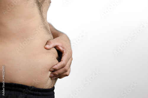 A side photo of a man with a hand on his protruding belly indicates fat and weight gain from unhealthy food. No exercise. Topless body. White background.