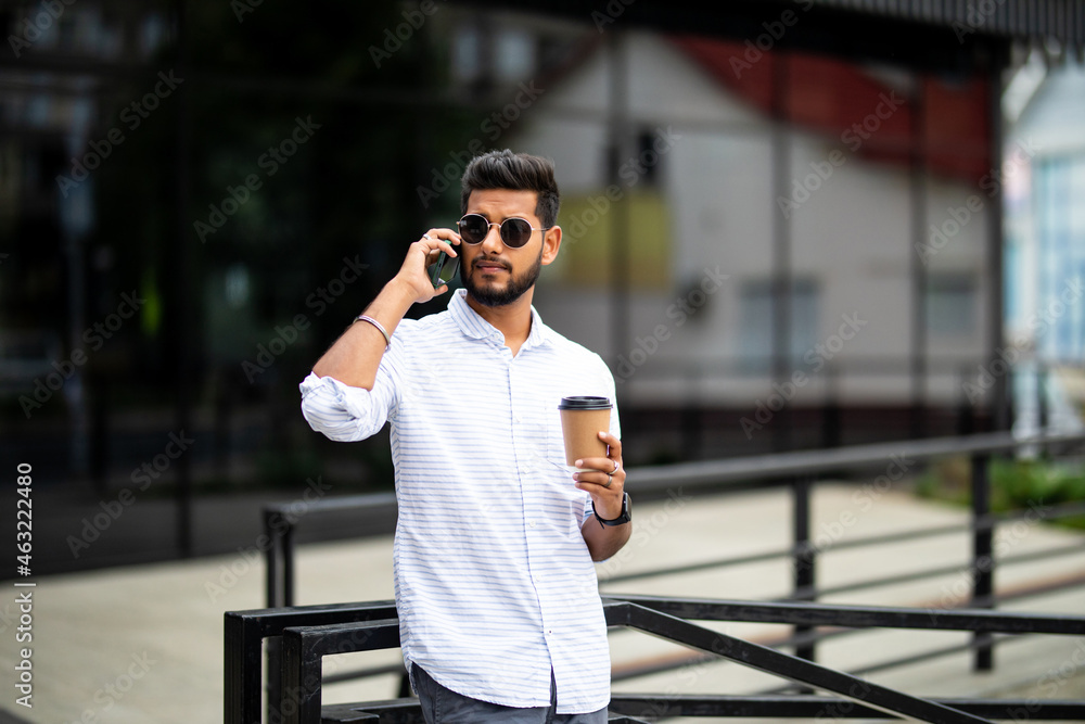 Business talk on a go. Confident young indian man in glasses holding coffee cup and talking on mobile phone while walking outdoors Business talk on a go. Confident young indian man in glasses holding coffee cup and talking on mobile phone while walking outdoors
