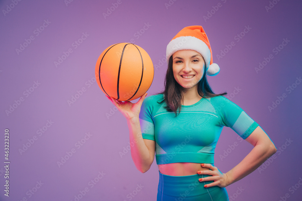 Fashion portrait of young fit and sportive caucasian woman in Christmas hat with basketball on gradient background. Perfect body ready for summertime. Beauty, resort, sport concept