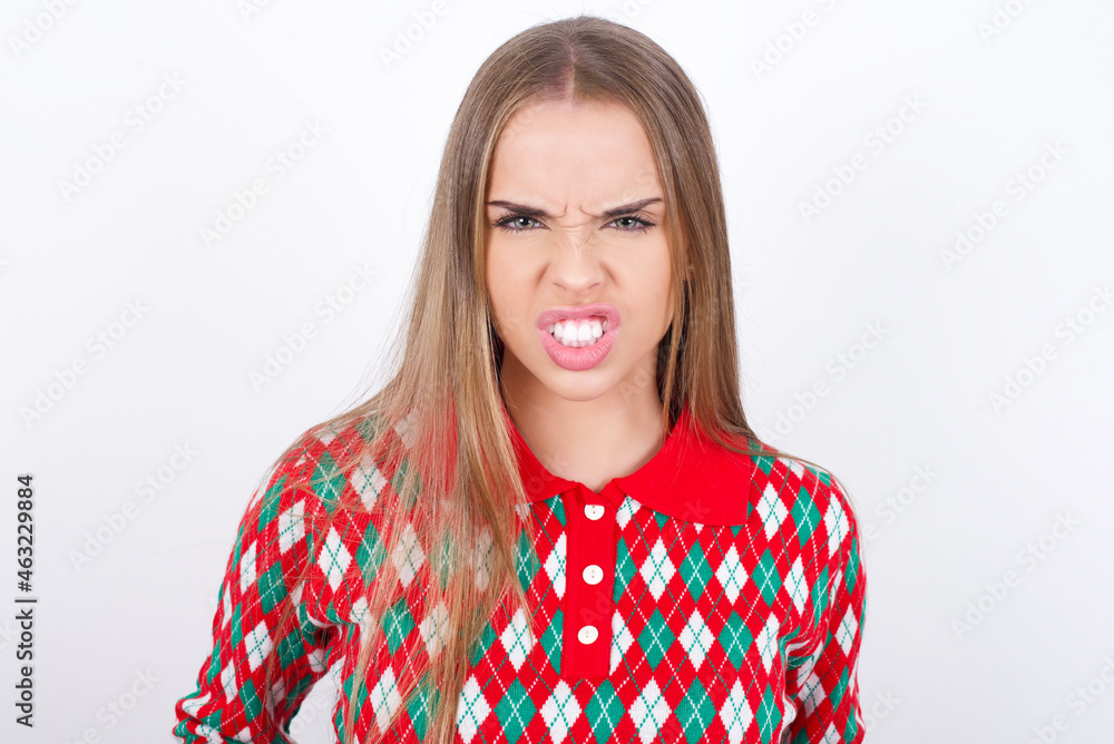 Mad crazy Young caucasian girl wearing christmas sweaters on white background clenches teeth angrily, being annoyed with coming noise. Negative feeling concept. Mad crazy Young caucasian girl wearing christmas sweaters on white background clenches teeth angrily, being annoyed with coming noise. Negative feeling concept.