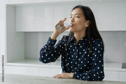 Young Asian woman drinks water from a glass sitting in the kitchen. A young woman smiles while holding a glass of water at home. Lifestyle concept.