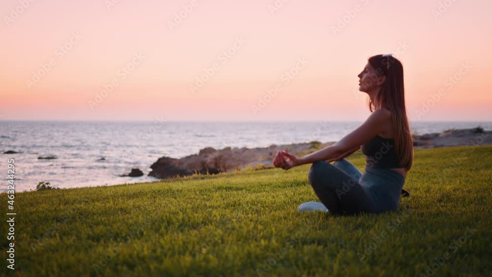 Side view of peaceful caucasian woman in activewear sitting on lotus position on beach grass and meditating with closed eyes. Yoga practises near ocean during evening time. Side view of peaceful caucasian woman in activewear sitting on lotus position on beach grass and meditating with closed eyes. Yoga practises near ocean during evening time.