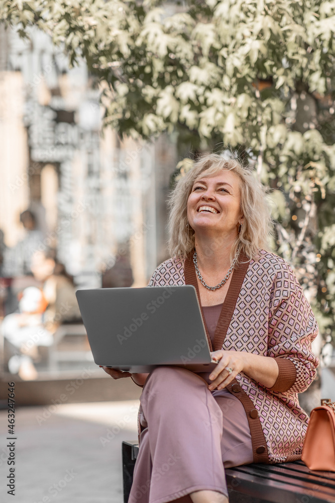 Attractive adult blonde of European appearance sitting on a bench in the city leads a video conference online with a laptop in her hands Attractive adult blonde of European appearance sitting on a bench in the city leads a video conference online with a laptop in her hands