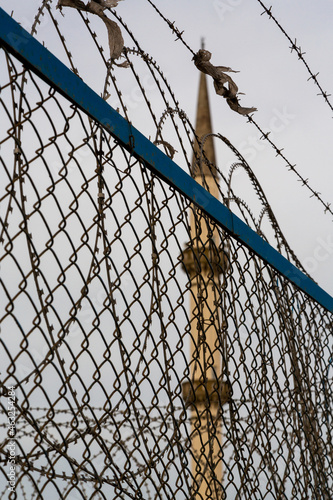 Concept of anti-Islam  Islamophobia  Wire mesh and barbed wire with a minaret in the background. The minaret is out of focus.