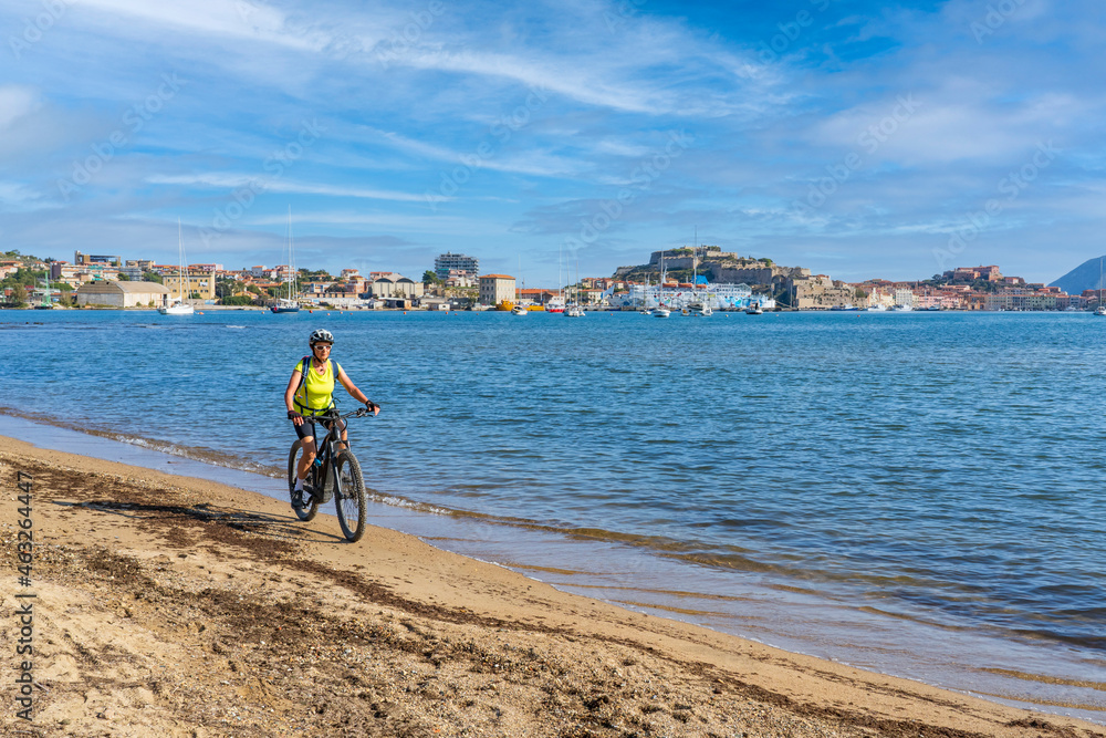 Fototapeta premium nice woman riding her electric mountain bike at the coastline of mediterranean sea on the Island of Elba in the tuscan Archipelago, in front of Porto Ferraio,Tuscany, Italy