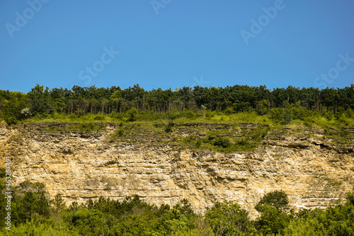 Cliff of ancient calcareous rocks, overgrown with forest, against blue sky. Sedimentary Cretaceous rocks of seabed. Horizontal photo. Copy space. Selective focus.