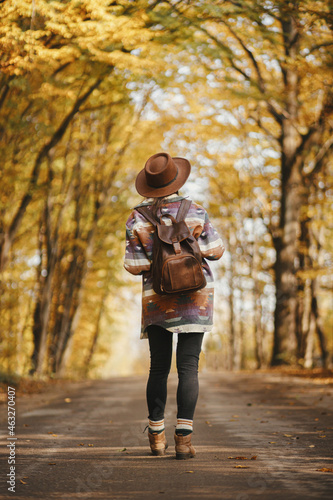 Stylish woman hipster in hat with backpack walking on road in sunny autumn woods. Young female traveler hiking in fall forest, beautiful moment. Travel and wanderlust concept. Back view © sonyachny
