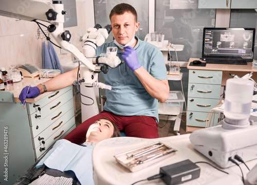 Handsome male stomatologist holding dental mirror while female patient lying in dental chair. Young man dentist sitting beside woman in dental office with modern equipment. Concept of dentistry.