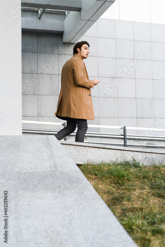 Side view of young businessman in brown coat holding smartphone while walking near building on street 