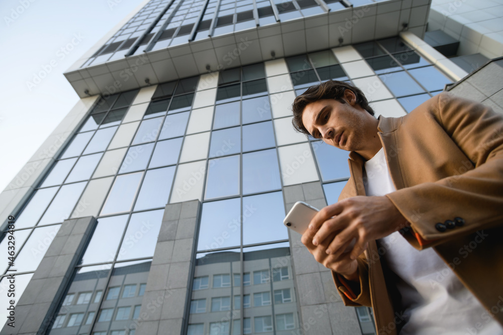 Fototapeta premium Bottom view of good looking businessman in autumn coat using cellphone near business center 