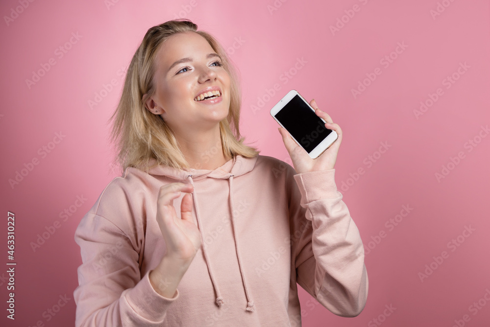 laughing young woman shows the phone screen and smiles . An attractive young woman in a pink hoodie uses a smartphone, a photo on pink. laughing young woman shows the phone screen and smiles . An attractive young woman in a pink hoodie uses a smartphone, a photo on pink.