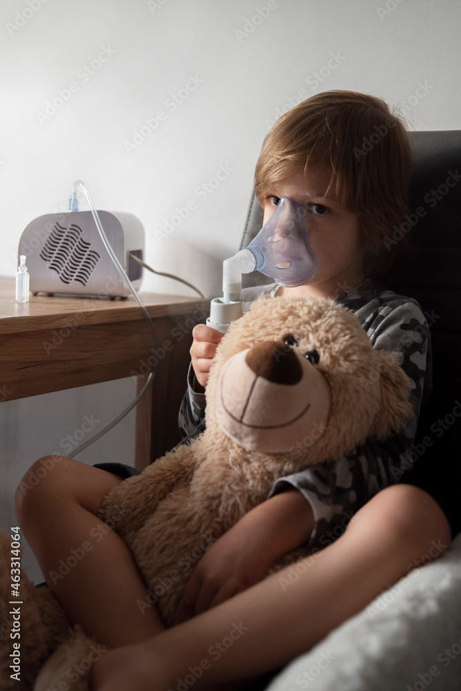Przeziębiony chłopiec siedzący przy stole z inhalatorem i pluszowym misiem - obrazy, fototapety, plakaty 