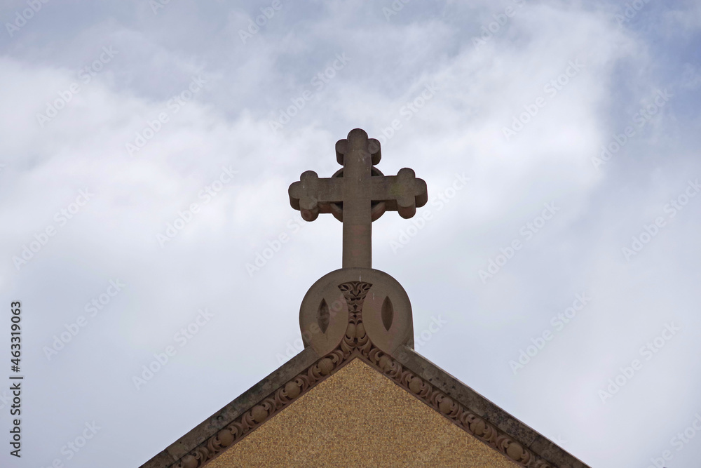 A decorative stone cross on the rooftop of a church with cloudy sky behind