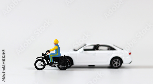 A miniature driver wearing a helmet next to a white miniature car and riding a motorcycle. Miniature people and miniature car.
