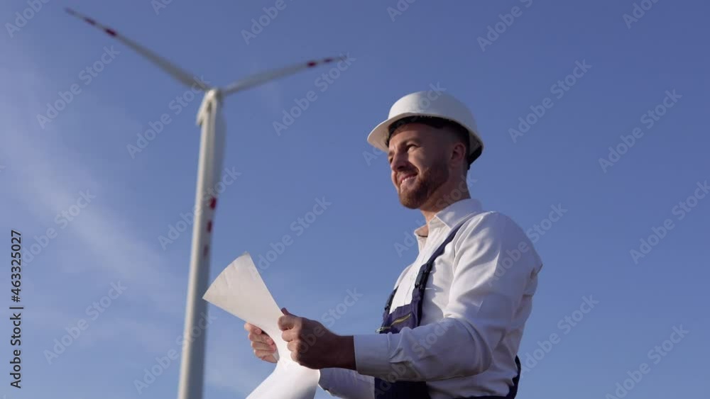 A male engineer in a white helmet and a classic shirt inspects the power plant's capacity and reads project drawings on a large sheet of paper