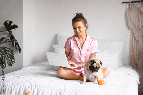 Young woman with cute pug dog using laptop in bedroom