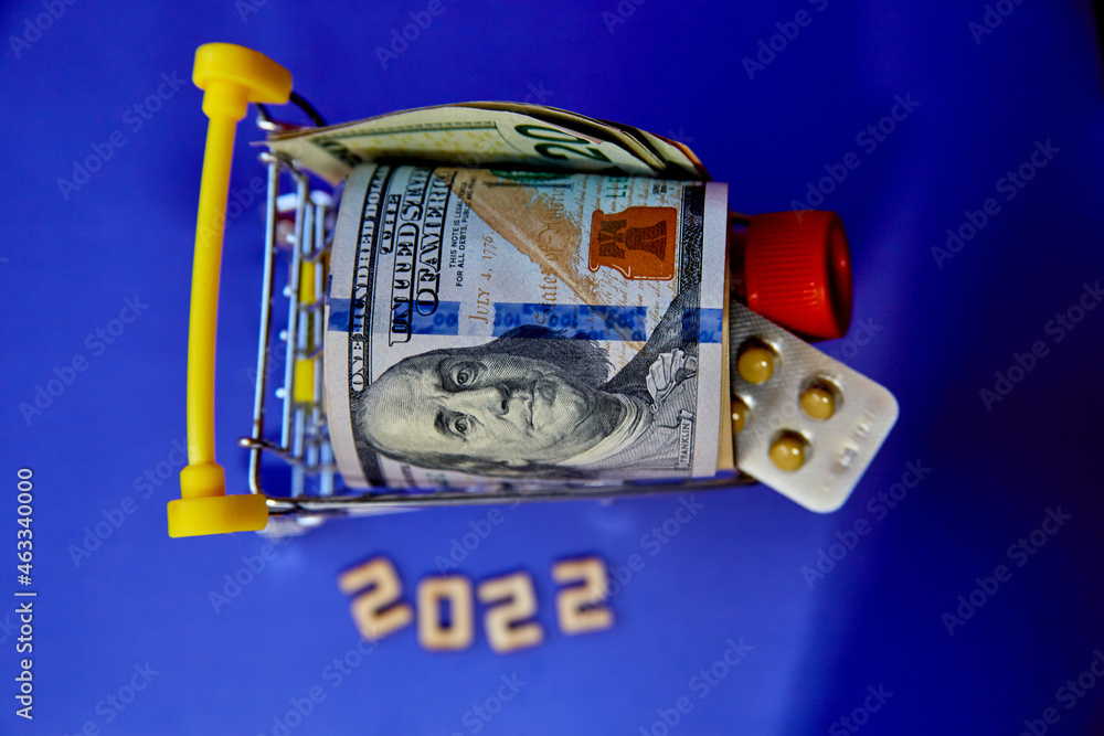 Fototapeta premium concept -Inscription 2022 and Shopping Cart Trolley with curled dollar banknotes and with a plate of pills and a medical tube against the purple background