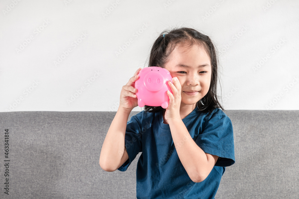 Young happy Asian little child girl holding the Piggy Bank for listing the coin inside and Sitting On Sofa At Home in the morning. Studio shot Copy Space. Personal saving money for the future concept Young happy Asian little child girl holding the Piggy Bank for listing the coin inside and Sitting On Sofa At Home in the morning. Studio shot Copy Space. Personal saving money for the future concept