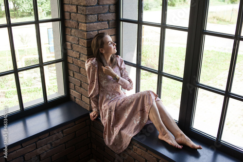 Beautiful young woman in pink dress resting on the windowsill barefoot. Happy smiling girl sits on the stained glass window at industrial loft wooden brick style interior. Leisure concept.