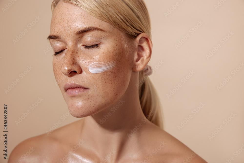 Fototapeta premium Close up of a calm and beaautiful blond woman with facial cream on a cheek against pastel background