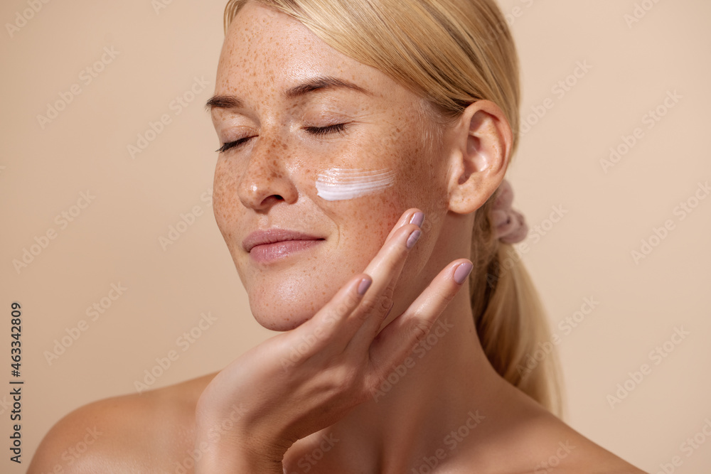 Close up of beautiful young female with cream on a face. Smiling woman with closed eyes touching cheek with fingers. - obrazy, fototapety, plakaty 
