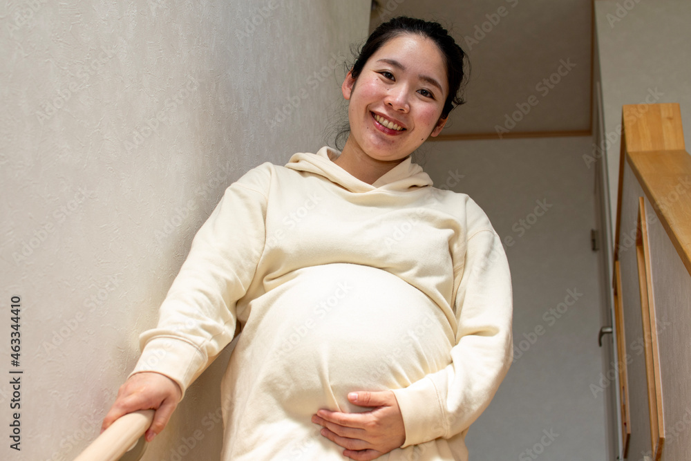 Obraz premium A young pregnant Asian woman with smile goes down the stairs carefully by using handrails in a house