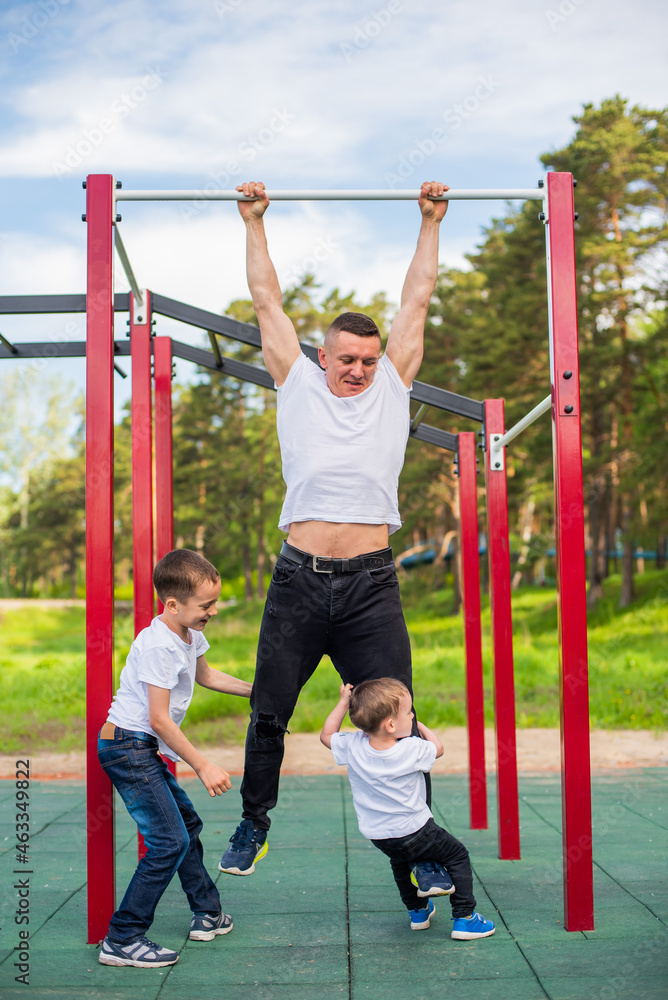 Obraz premium Caucasian man and two boys doing exercises outdoors. The father pulls himself up on the horizontal bar with his sons on the playground.