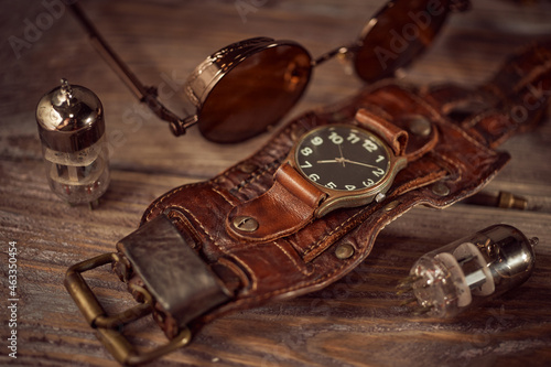 Steampunk still life - old vintage objects on a wooden background. Leather wrist watch, dark glasses, old tube lamps, magnifying glass, gears