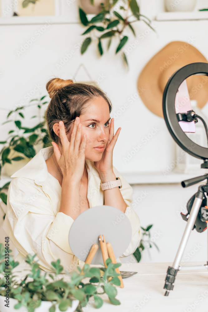 A young woman beauty blogger broadcasts live streaming to review makeup and cosmetic products on social media,reviews beauty product for video blog.Influencer uses smartphone on tripod and ring lamp.
