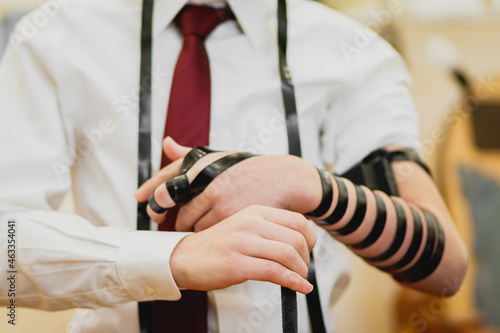  A close-up of a Jewish bar mitzvah boy, wearing a white shirt and a red tie. wearing a tefillin for the first time in a synagogue photo