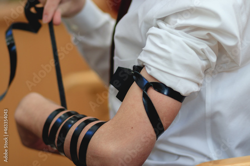  A close-up of a Jewish bar mitzvah boy, wearing a white shirt, put s tefillin for the first time in a synagogue photo