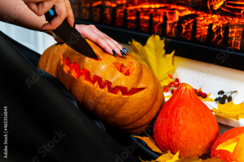a witch in a raincoat with a knife carves a pumpkin for Halloween at home, against the background of the fireplace